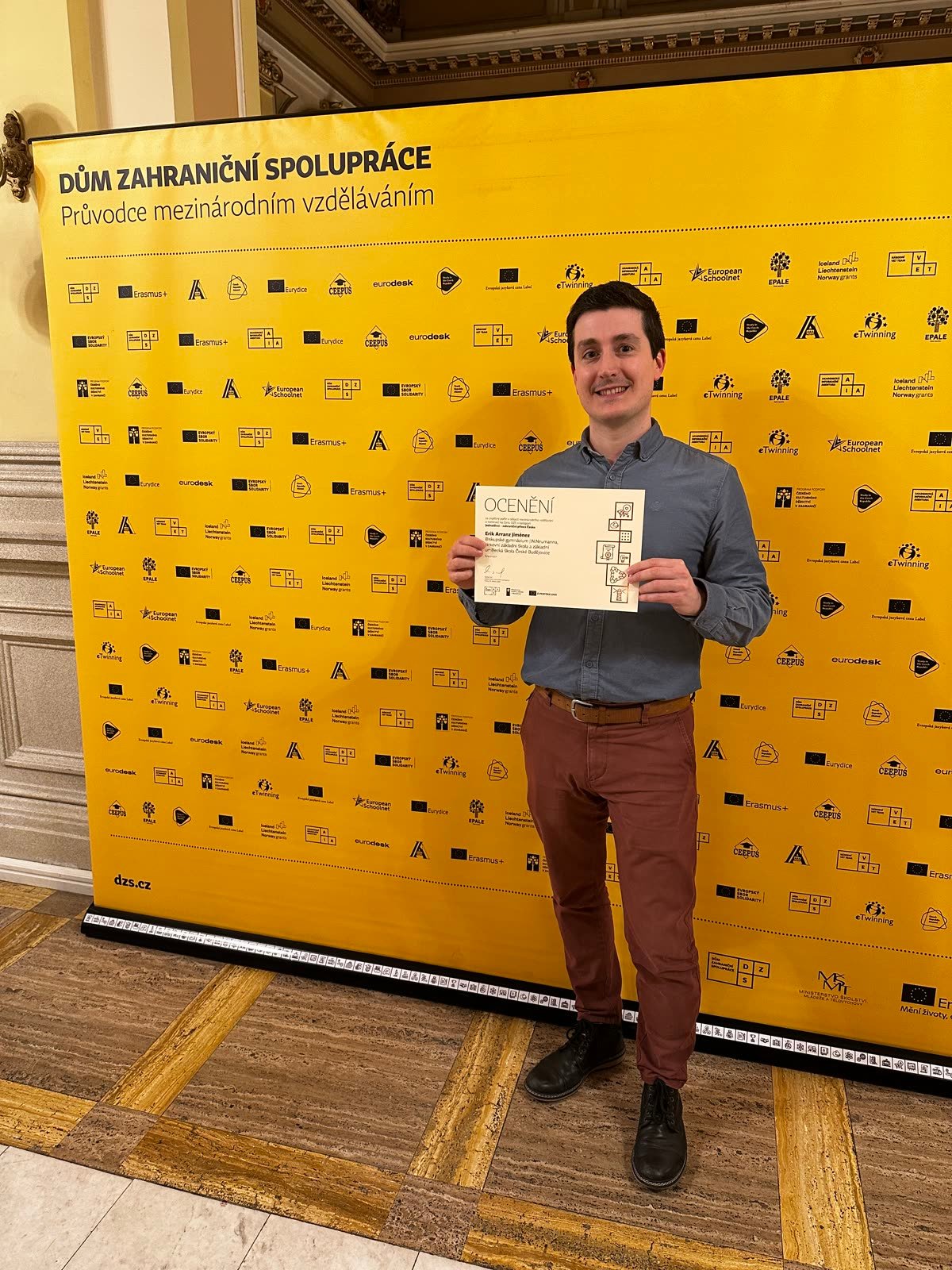 Erik Arranz Jiménez con el diploma de nominación delante del photocall amarillo del DZS en la gala de Praga